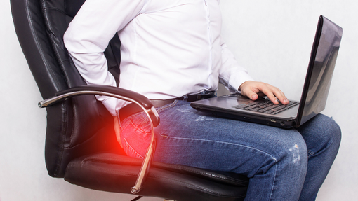 man sitting on a chair with a laptop clutching his buttocks due to sciatica pain